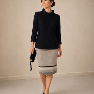 Op de afbeelding: Een vrouw draagt een zwarte jas met een hoge kraag en een grijze potloodrok. De rok heeft horizontale strepen in grijs en zwart. Ze houdt een zwarte clutch vast en draagt zwarte hakken. Formele outfit.