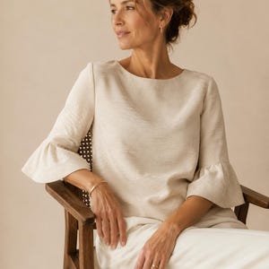 Op de afbeelding: Een crèmekleurige blouse met driekwart mouwen en uitlopende manchetten. De blouse heeft een ronde halslijn en wordt gecombineerd met een witte broek. De persoon zit op een houten stoel met een geweven zitting.