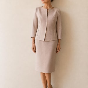 Op de afbeelding: Lichtbeige rokpak. De jas heeft een getailleerde pasvorm met driekwartmouwen. De knielange rok vult de jas aan. Beige pumps en een parelketting maken de outfit compleet. Het pak is gemaakt van een stof met textuur.
