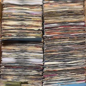 May include: A large cardboard box filled with a dense collection of vintage vinyl records stored vertically. The records are organized with blue and green paper dividers, some featuring handwritten labels like 80s and 180-189, creating a structured archive of music media.