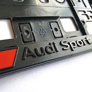 Può includere: Cornice per targa in plastica nera con scritta argentata "Audi Sport". Un emblema rosso e nero a forma di rombo è visibile a sinistra. La cornice presenta aperture rettangolari e fori di montaggio.