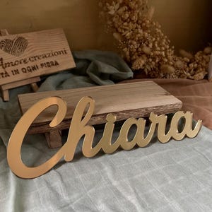 May include: A gold-colored, reflective acrylic name sign featuring the name Chiara in a cursive script font. The sign is positioned in front of a small wooden block, with a wooden plaque displaying the text Amore creazioni TÀ IN OGNI FIBRA.