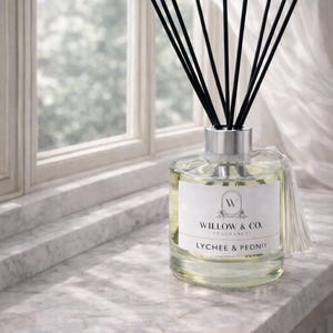 May include: A glass diffuser bottle with black reeds and a silver cap, labelled "Willow & Co. Fragrances, Lychee & Peony." The bottle is on a marble windowsill, with a white tassel and a window in the background.