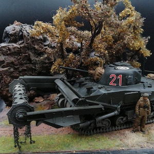 May include: A detailed model of a dark green tank with the number 21 in red on the turret. The tank has a blade attachment and is positioned in a diorama with a rock, tree, and a miniature figure of a soldier.