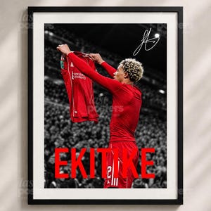 May include: A framed sports poster featuring a football player with blonde curly hair wearing a red long-sleeve jersey and shorts, holding up a red jersey. The name EKITIKE is printed in large red letters at the bottom, with a white signature above.