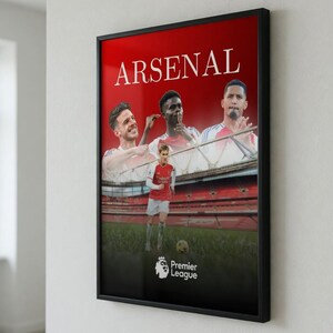 May include: Framed poster featuring the word "ARSENAL" in white on a red background. The poster showcases images of Arsenal football players, with the Premier League logo at the bottom. The poster is displayed on a white wall.