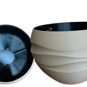 May include: A modern, abstract decorative bowl with a unique design. The bowl is light beige with a black interior and a circular lid. The bowl's exterior features a layered, wave-like pattern. The lid is black with a circular design.