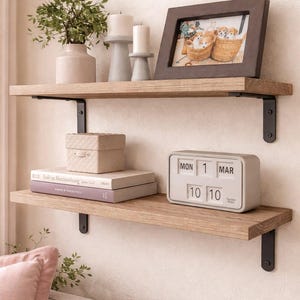 Puede incluir: Dos estantes de madera con soportes metálicos negros están montados en una pared beige. El estante superior contiene un marco de fotos, velas y un jarrón con vegetación. El estante inferior muestra libros, una caja y un calendario.