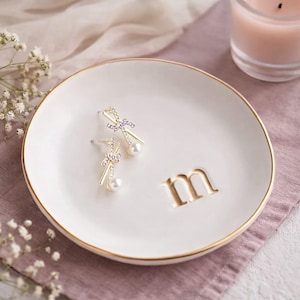 Könnte beinhalten: Eine weiße Keramik-Schmuckschale mit goldenem Rand und dem Buchstaben "m" in Gold. Zwei goldene Ohrringe mit lila Akzenten und Perltropfen liegen auf der Schale. Eine brennende Kerze und Blumen sind im Hintergrund.