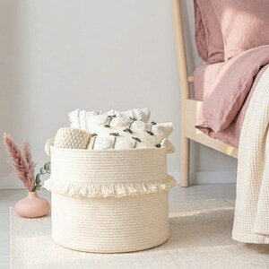 May include: A cream-colored, round woven storage basket with a fringed trim. The basket is filled with white pillows and a small woven container. A pink vase with dried flowers and a bed with pink bedding are in the background.