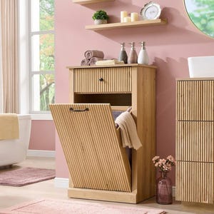 May include: A light wood laundry hamper cabinet featuring a top drawer with a single black knob and a tilt-out front panel with a black handle. The cabinet has a vertical ribbed texture on the drawer and front panel, with an open interior compartment.