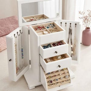May include: A white jewelry armoire with a mirrored top and multiple drawers and compartments. The armoire is filled with various jewelry pieces, including necklaces, bracelets, rings, and a watch. The armoire has a classic design and is made of wood.