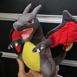 May include: A plush Charizard toy in shades of gray, red, and yellow. The toy has large wings, a yellow belly, and white claws. The mouth is open, revealing a pink tongue and teeth. The toy is held in a person's hand.