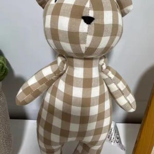 Puede incluir: Un oso de peluche con un estampado de cuadros marrones y blancos. El oso tiene una nariz negra y orejas pequeñas. El juguete está hecho de tela suave y está diseñado para niños.