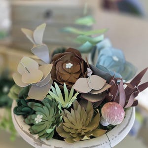 Op de afbeelding: Een witte, verweerde keramische plantenbak gevuld met papieren bloemen in tinten groen, bruin en blauw. Het arrangement bevat verschillende succulenten en bloemmotieven, met kleine witte accenten. Een decoratief stuk voor de woondecoratie.