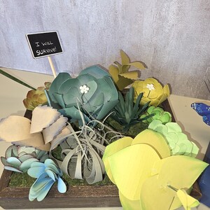 Op de afbeelding: Een houten plantenbak gevuld met papieren vetplanten in groen, blauw en geel. Een klein bordje leest "I WILL SURVIVE!". De opstelling bevat verschillende papieren plantontwerpen.