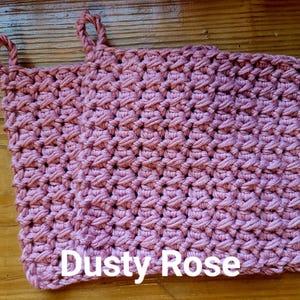 May include: A dusty rose-colored, crocheted potholder with two looped handles. The handmade kitchen accessory features a textured stitch pattern and the text "Dusty Rose" in white block letters.
