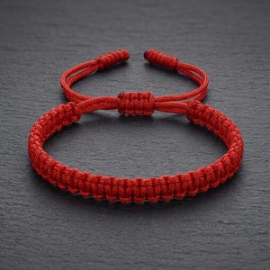May include: A vibrant red macrame bracelet with an adjustable sliding knot closure. The bracelet is made of woven cord, showcasing a detailed texture. The design includes two adjustable cords and a central knot, all in a matching bright red color.