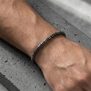May include: A dark gray beaded bracelet. The bracelet is made of small, cylindrical beads strung together. The bracelet is worn on a wrist, with the skin tone of the person wearing the bracelet visible.