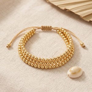 Puede incluir: Una pulsera de tres hileras con pequeñas cuentas metálicas doradas y pulidas dispuestas en tres filas paralelas. La pulsera incluye un cierre de cordón beige ajustable con dos pequeñas cuentas doradas en los extremos y una concha de cauri blanca.