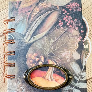 Puede incluir: Un cuaderno con espiral con una cubierta de estilo vintage que presenta un conejo, hojas y una calabaza en un marco ovalado. La paleta de colores incluye tonos de marrón, verde y naranja. El cuaderno tiene una encuadernación en espiral color cobre.