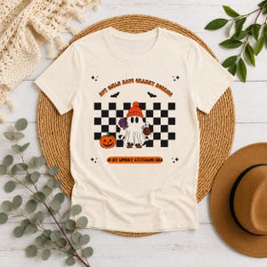 May include: A cream-coloured short-sleeve t-shirt featuring a graphic of a ghost wearing an orange beanie, holding a ball of yarn and a needle. The design includes a black and white checkerboard background, a pumpkin, bats, and the text "HOT GIRLS HAVE GRANNY HOBBIES" and "IN MY SPOOKY STITCHING ERA."