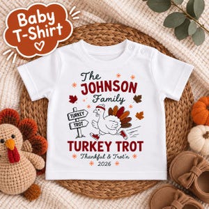 May include: A white short-sleeved baby t-shirt featuring a graphic of a running turkey wearing a headband and sneakers. The text reads The Johnson Family, Turkey Trot, Thankful & Trot'n, and 2026, surrounded by autumn leaves.