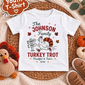May include: A white short-sleeved youth t-shirt featuring a graphic of a turkey wearing a Santa hat and sneakers. The text reads The JOHNSON Family TURKEY TROT Thankful & Trot'n 2026, accompanied by a signpost, autumn leaves, and star accents.