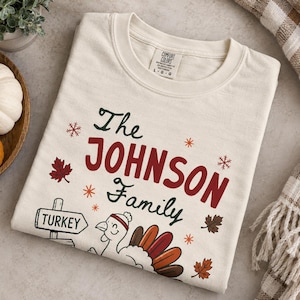 May include: A cream-colored t-shirt featuring the text The JOHNSON Family in dark green and red. The graphic includes a turkey wearing a hat, a signpost labeled TURKEY, and various autumn leaves and snowflake icons printed on the front.