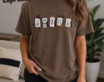 May include: An espresso-colored t-shirt featuring a graphic of five anthropomorphic mahjong tiles dressed in Halloween costumes, including a witch, a vampire, a pumpkin, a bat, and a devil, with the word Espresso printed in the top left corner.
