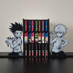 Op de afbeelding: Zwart-witte manga boekensteunen met anime-personages. Tussen hen in staan zeven manga boeken met kleurrijke ruggen, getiteld "Yomi no Tsugai" en genummerd van 1 tot en met 7. De boeken staan in een rij.