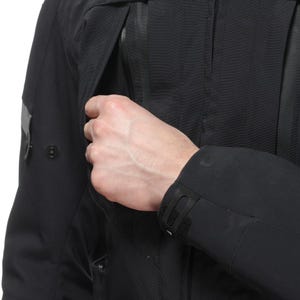 May include: A close-up of a black jacket with a zippered pocket being opened. The jacket has a textured appearance and a small logo on the sleeve. The cuff features a Velcro closure.