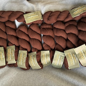 May include: Several skeins of brown yarn are arranged on a light-colored surface. Each skein is labeled with a tag that reads "CASCADE YARNS" and "CASCADE 220 Fingering". The yarn appears soft and is a rich, earthy tone.