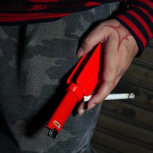 Könnte beinhalten: Ein rotes BIC-Feuerzeug mit einzigartigem Design wird in der Nähe einer brennenden Zigarette gehalten. Das Feuerzeug wird vor einer Person gehalten, die ein rot-schwarz gestreiftes Hemd und Camouflage-Hosen trägt. Das Feuerzeug ist rot mit dem BIC-Logo.