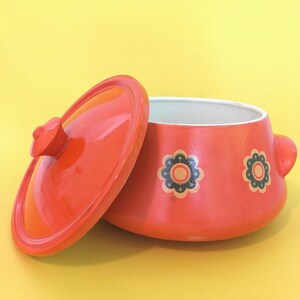 May include: A bright orange ceramic casserole dish with a matching lid. The exterior features a repeating pattern of stylized dark blue and cream-colored flowers. The interior of the pot is white, and the vessel includes two small side handles.