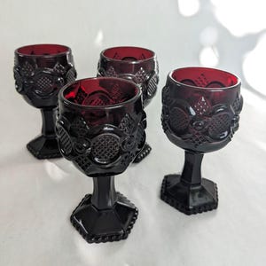 May include: A set of four deep red glass goblets featuring a textured, pressed pattern with circular and diamond motifs. Each goblet has a thick, hexagonal stem and a matching hexagonal base, showcasing a rich, dark crimson hue throughout the glass.