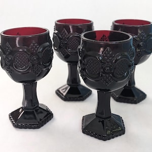 May include: A set of four deep red glass goblets featuring a raised geometric pattern on the bowl. Each goblet has a thick stem and a hexagonal base. The glass is translucent, showing a darker shade of red where the pattern is embossed.