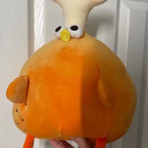 May include: A soft toy chicken with a large orange body, small orange wings, and orange legs. The chicken has a cream-coloured head and a yellow beak. The eyes are white with black pupils. The toy is designed to be cuddly.