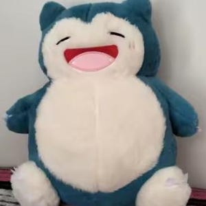 May include: A plush Snorlax toy in shades of blue and white. The toy has a large, round body, small arms and legs, and a smiling face with a pink tongue. The toy is soft and cuddly, perfect for fans of the popular franchise.