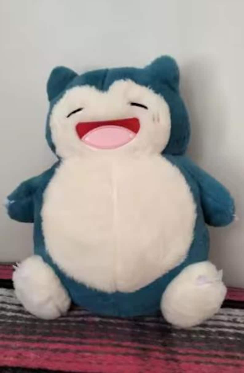 May include: A plush Snorlax toy in shades of blue and white. The toy has a large, round body, small arms and legs, and a smiling face with a pink tongue. The toy is soft and cuddly, perfect for fans of the popular franchise.