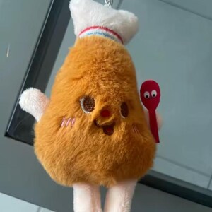 May include: A novelty keychain featuring a cartoon chicken drumstick character. The plush toy is brown with embroidered eyes, a nose, and a smiling mouth. It wears a chef's hat and holds a red spoon. The hat has a red and blue stripe.