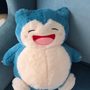 May include: A plush toy of a Snorlax character, featuring a light blue body, a cream-colored belly, and small arms and feet. The toy has a smiling face with closed eyes, a pink mouth, and rosy cheeks. It is sitting on a blue surface.