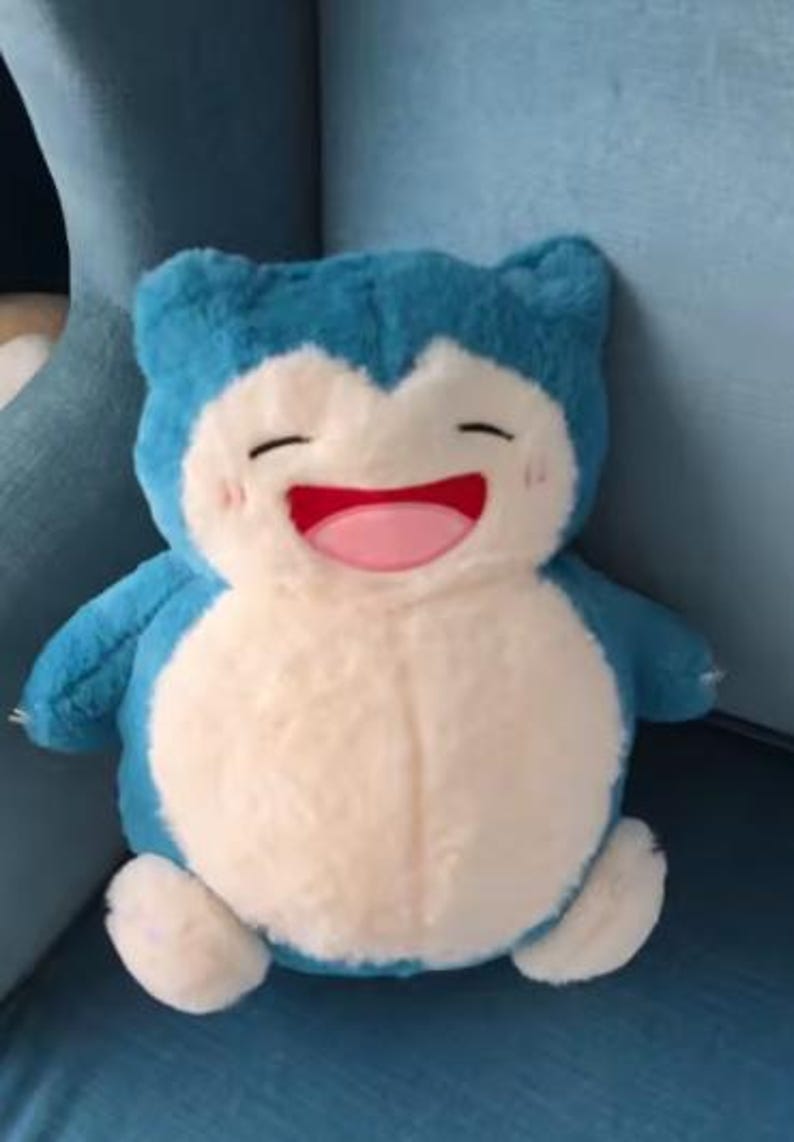 May include: A plush toy of a Snorlax character, featuring a light blue body, a cream-colored belly, and small arms and feet. The toy has a smiling face with closed eyes, a pink mouth, and rosy cheeks. It is sitting on a blue surface.