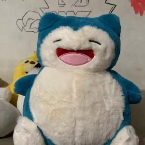 May include: A plush Snorlax toy, featuring a blue head and limbs, a white body, and a smiling face with a pink mouth. The toy is soft and cuddly, perfect for fans of the popular franchise.