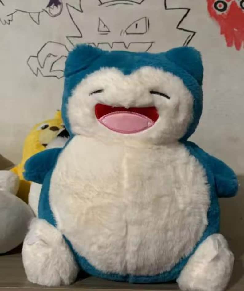 May include: A plush Snorlax toy, featuring a blue head and limbs, a white body, and a smiling face with a pink mouth. The toy is soft and cuddly, perfect for fans of the popular franchise.