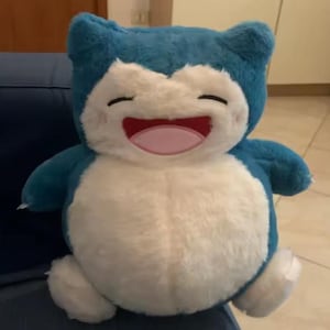 May include: A plush toy of a smiling, cartoon-like character. The toy has a blue head and limbs, a white belly, and a pink mouth. The toy is sitting on a blue surface.