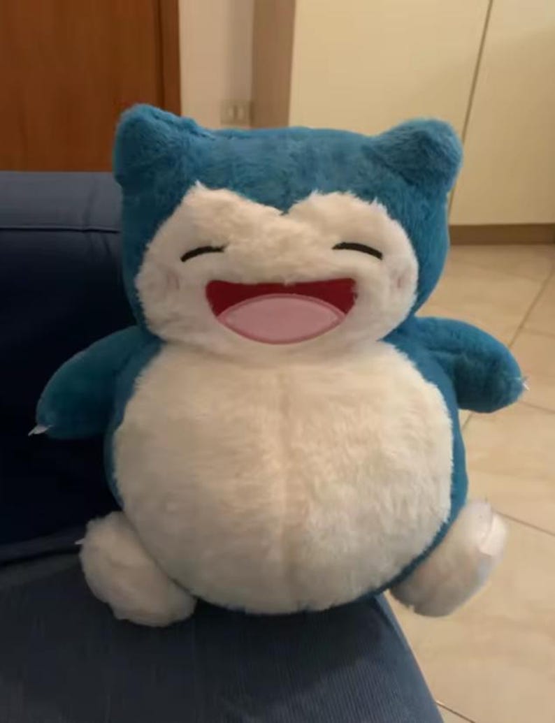May include: A plush toy of a smiling, cartoon-like character. The toy has a blue head and limbs, a white belly, and a pink mouth. The toy is sitting on a blue surface.