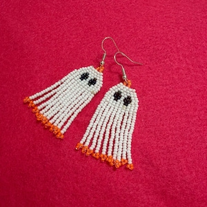 May include: A pair of ghost-shaped drop earrings crafted from white seed beads. Each earring features two black beads for eyes and a fringe of orange beads along the bottom edge, suspended from silver-toned metal earring hooks for pierced ears.