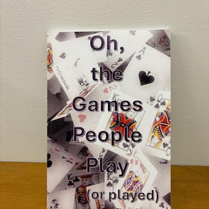 Può includere: Un libro in brossura intitolato Oh, the Games People Play (or played) Third Edition. La copertina presenta un collage di varie carte da gioco, incluse figure e un jolly, con il titolo stampato in un grande testo viola scuro senza grazie.
