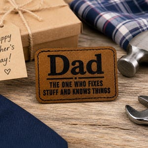 May include: A brown leather patch with the word "Dad" in black, and the text "THE ONE WHO FIXES STUFF AND KNOWS THINGS". A gift box with a "Happy Father's Day!" tag, a hammer, and a wrench are also visible.